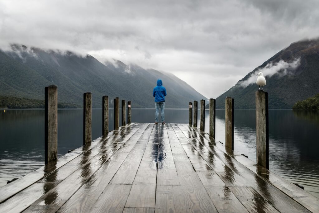 Person in blue jacket stands on a rainy dock overlooking serene lake and mountains.